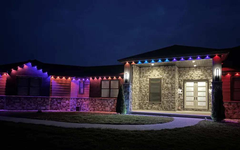 Stone front entry and ranch roofline lit in blue, purple, and amber permanent lighting at night