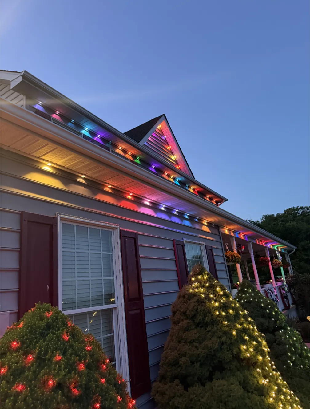 Gabled roofline and porch lit in a rainbow color scene with holiday lights on the front shrubs